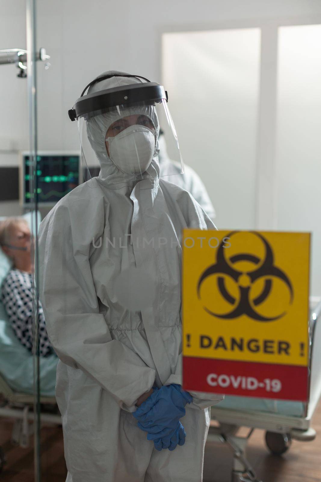 Nurse with face mask and coverall in hospital room, looking tired at camera, during coronavirus outbreak, to prevent infection. Patient laying in bed getting intravenous medicine treatment.