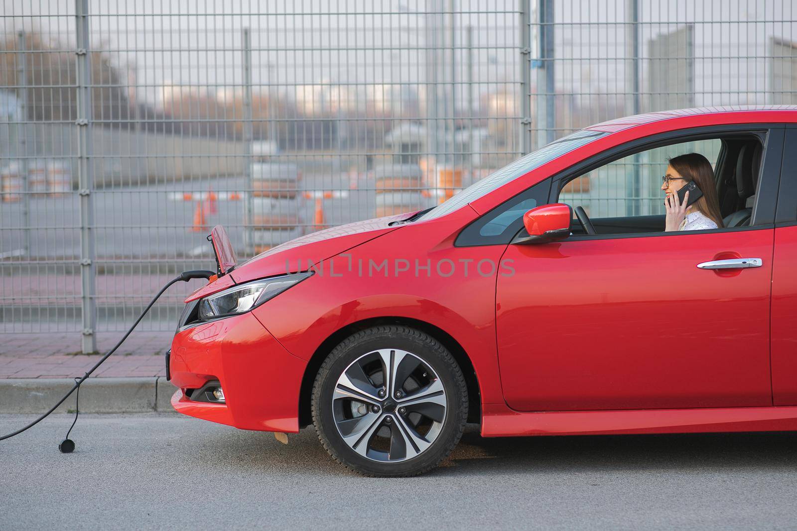 Woman using smart phone and waiting power supply connect to electric vehicles for charging the battery in car. Positive young girl talking on the phone sits in electric car and charging