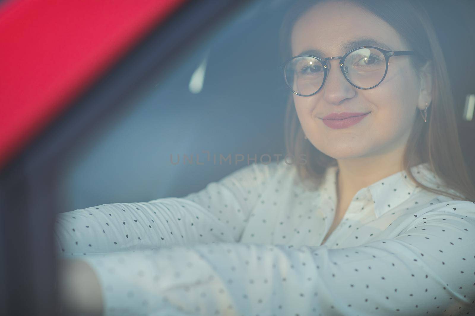 Portrait of a girl in glasses through the window of a car. Positive looking girl. Beautiful girl sitting in the new red car