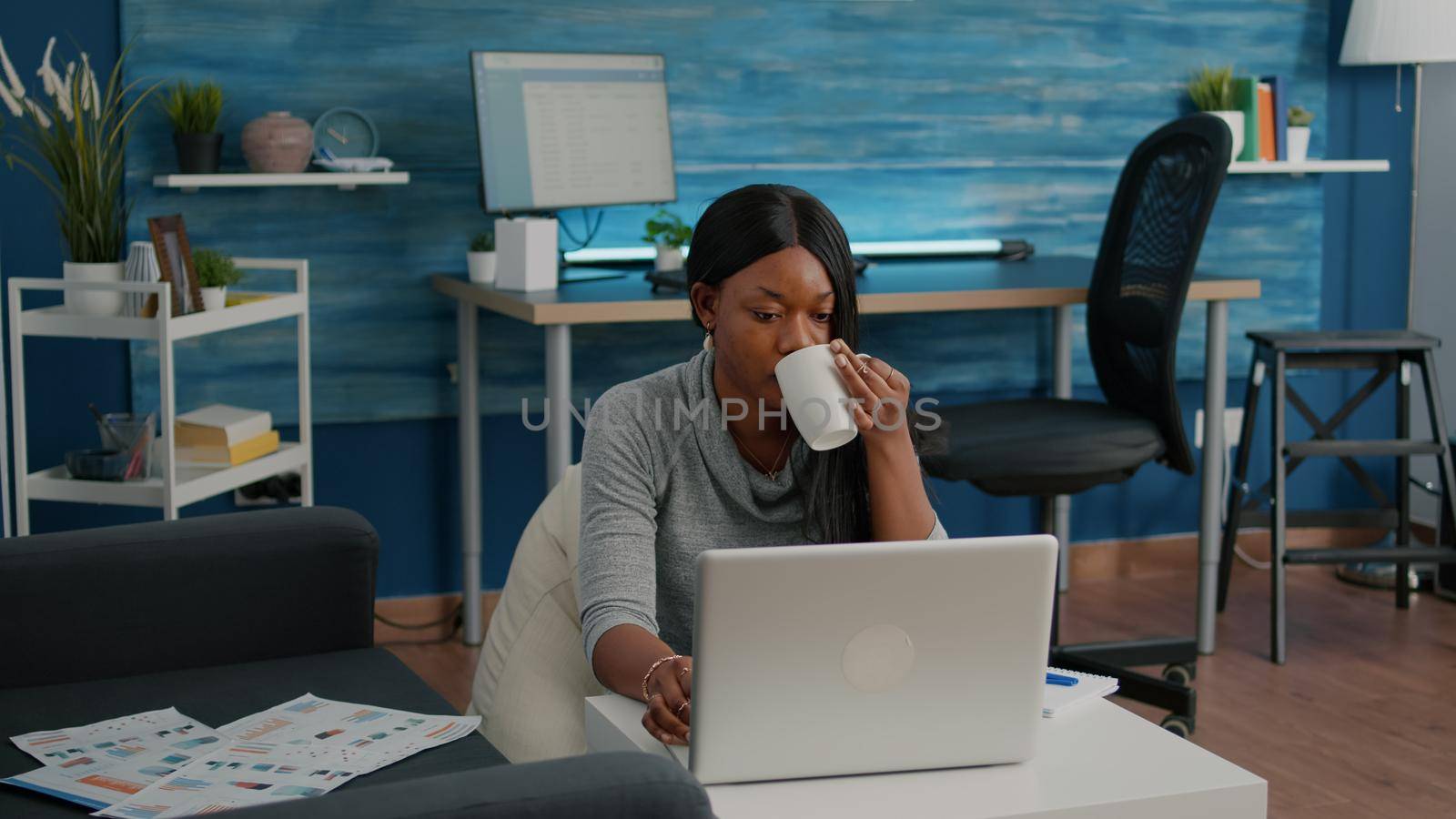 Black student drinking coffee typing social media article browsing lecture communication webinar on laptop working in living room. Young woman solving problem typing education email