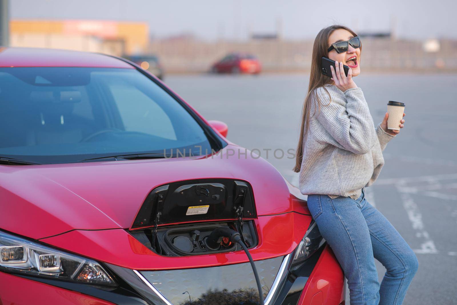 Girl Use Coffee Drink While Using Smart Phone and Waiting Power Supply Connect to Electric Vehicles for Charging the Battery in Car by uflypro