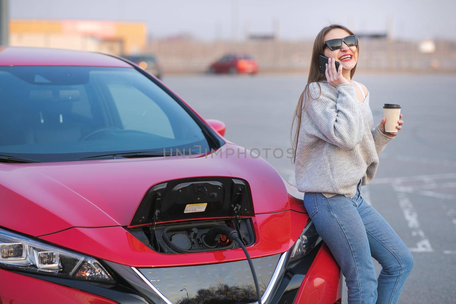 Girl Use Coffee Drink While Using Smart Phone and Waiting Power Supply Connect to Electric Vehicles for Charging the Battery in Car.