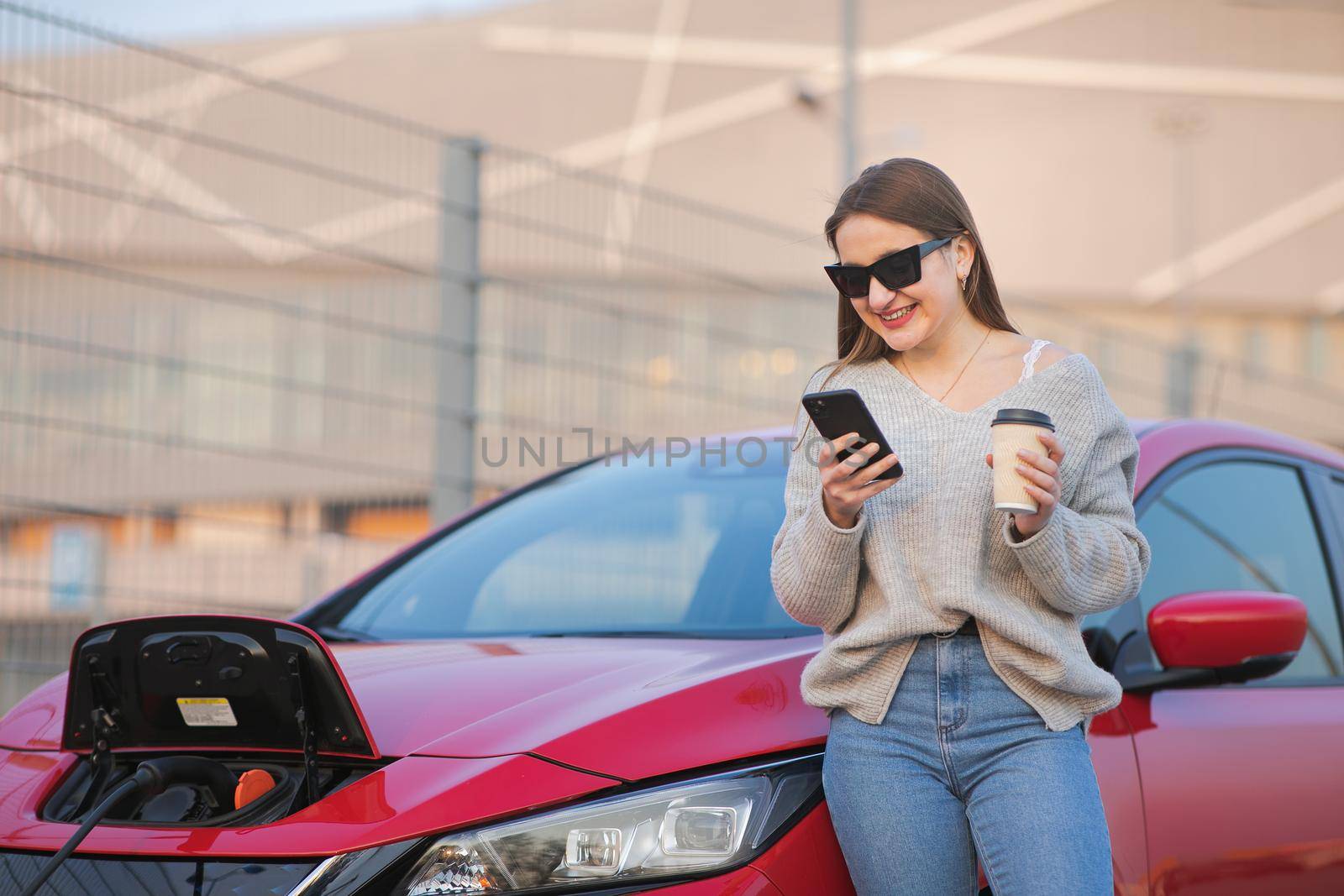 Woman stands with phone near her electric car and waits when vehicle will charged. Charging of electric car by uflypro