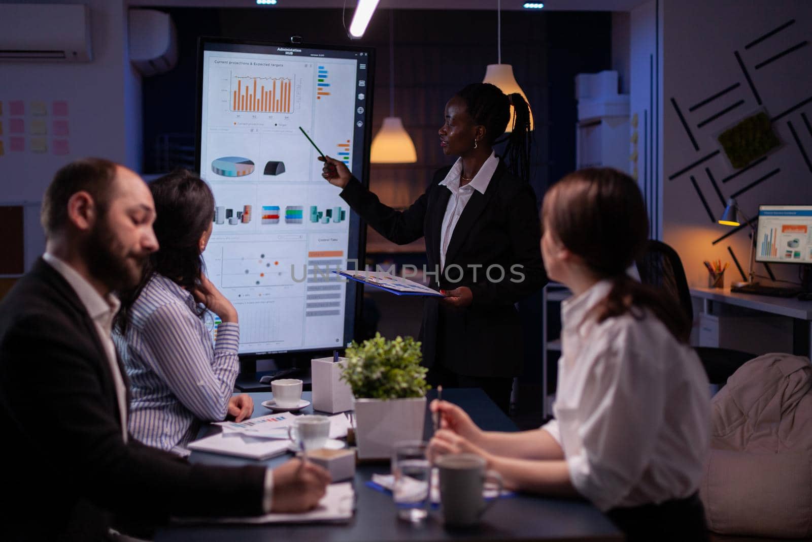 Workaholic african american leader standing in front of presentation monitor explaining marketing project solution late at night in company meeting room. Diverse teamwork working at business ideas.