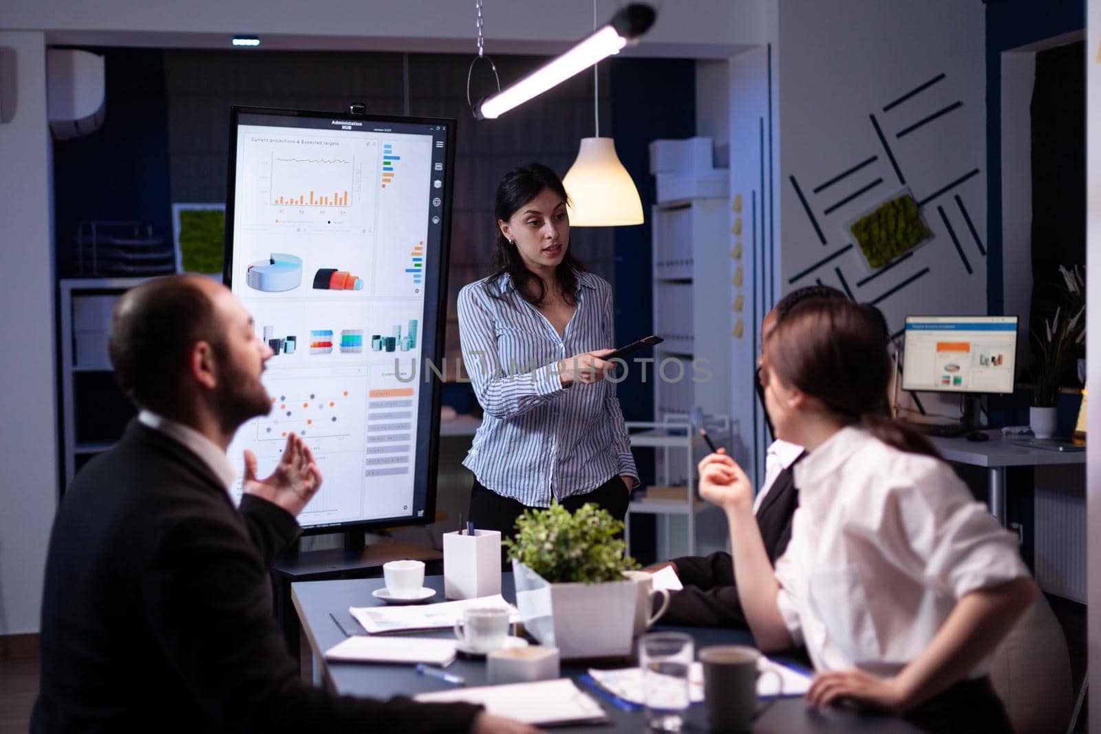 Workaholic businesswoman pointing financial strategy using monitor working overtime in company meeting office room. Diverse multi-ethnic teamwork overworked solving management statistics in evening