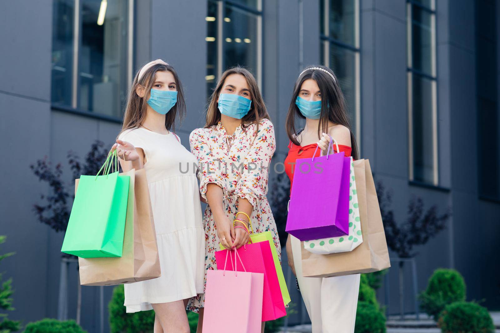 Group of young beautiful women in casual dresses, top and pants wearing masks to protect coronavirus pandemic standing in front of the shopping mall with colored bags in hands.