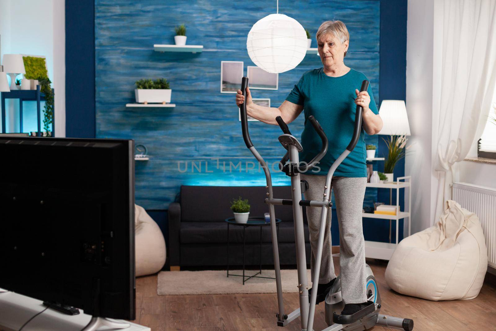 Invalid senior woman doing aerobics on cycling bike machine in living room for well being by DCStudio