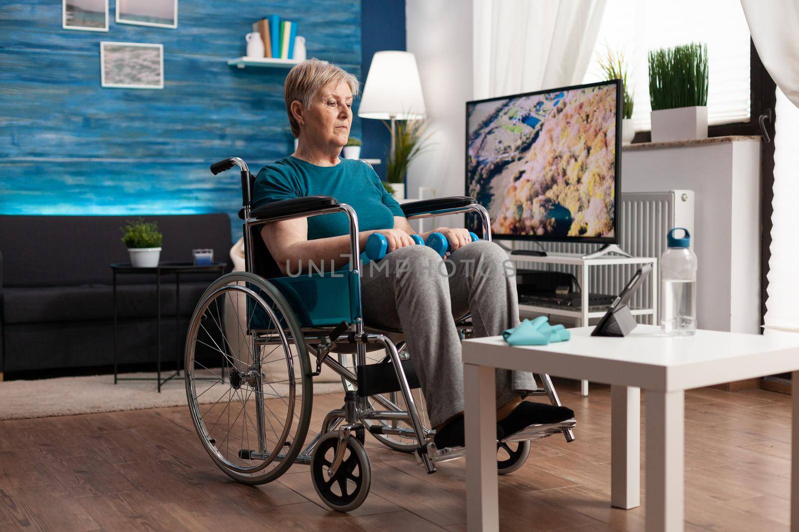 Disability senior woman in wheelchair looking at gymnastic online video on tablet computer by DCStudio