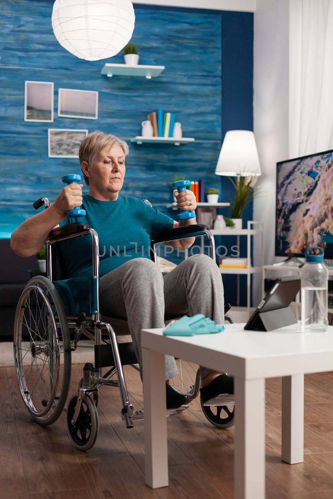 Handicapped senior woman holding trainer dumbbells doing arms exercise working at body persistence training in living room. Disabled pensioner looking at online muscles workout course on tablet