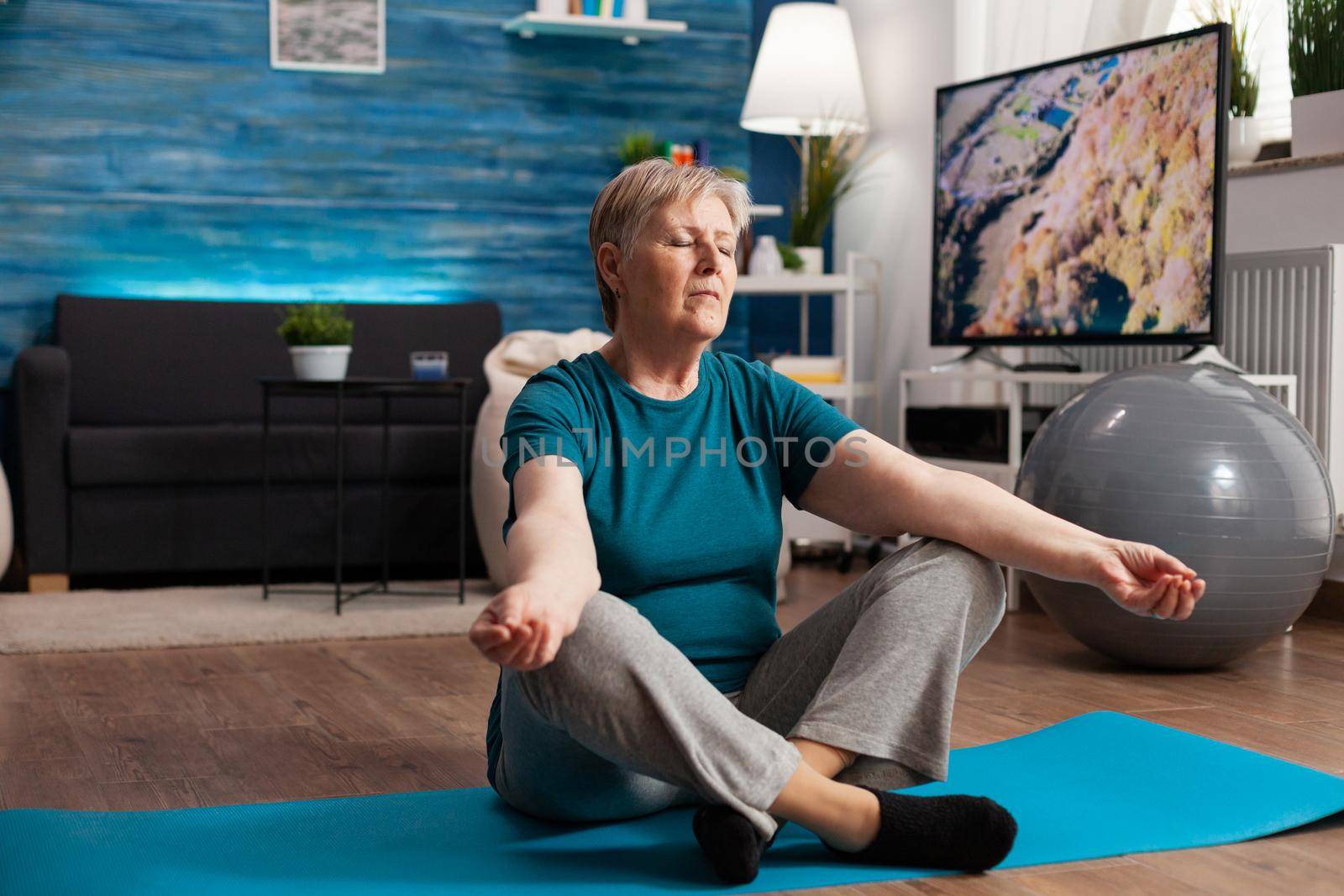 Peaceful senior woman with closed eyes sitting on yoga mat meditating during wellness workout by DCStudio