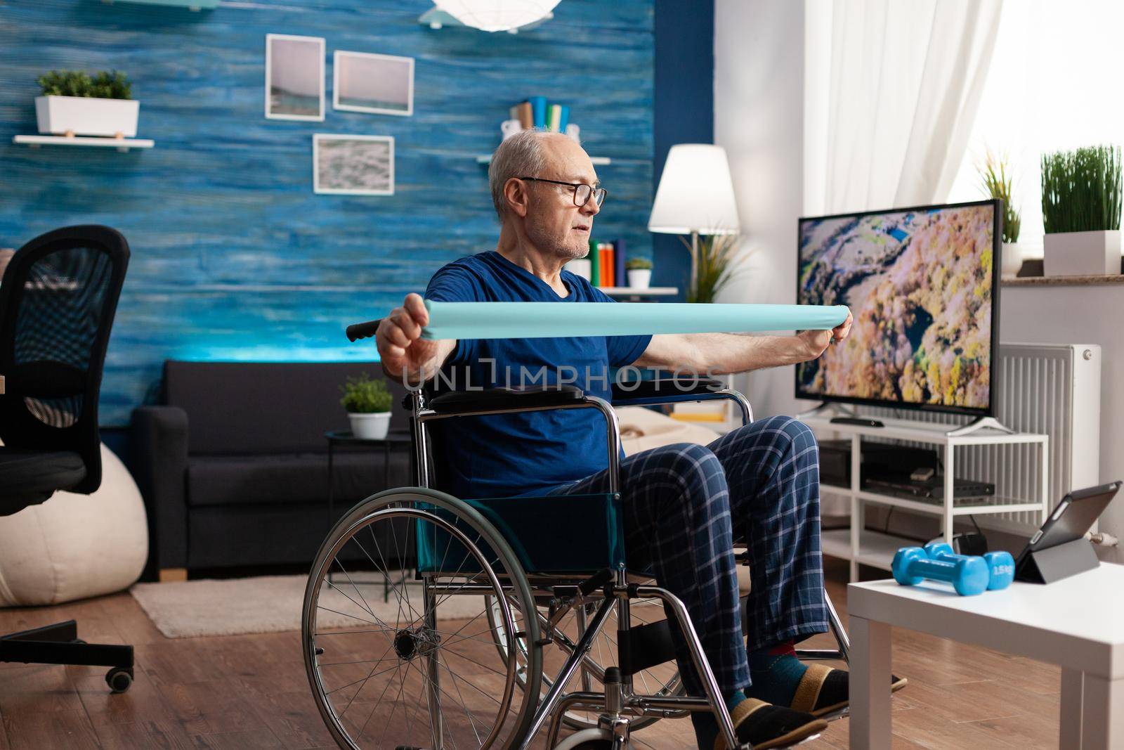 Handicapped old man in wheelchair training arm resistance exercising body muscle using elastic band. Pensioner doing gym workout after suffering accident watching healthcare therapy video on tablet