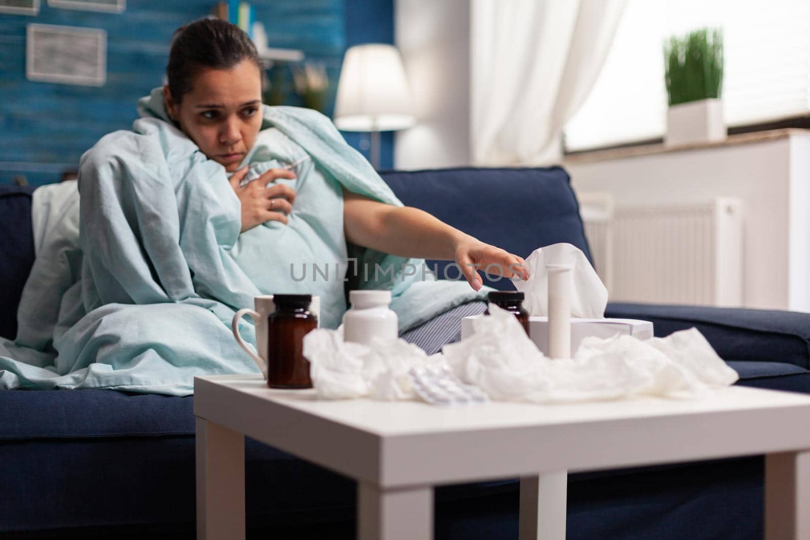 Sick woman in blanket sitting at home with coronavirus symptoms of temperature fever headache. Caucasian adult with medicine against flu illness infection disease wrapped in blanket