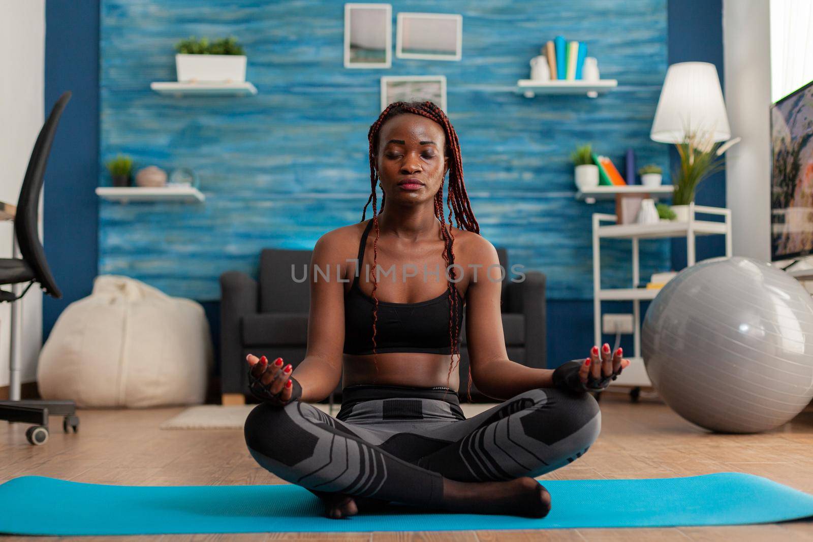 Calm tranquil black woman keeping eyes closed sitting on yoga mat, spending time on spirituality healthy lifestyle. Practicing meditating in lotus pose in home living room.