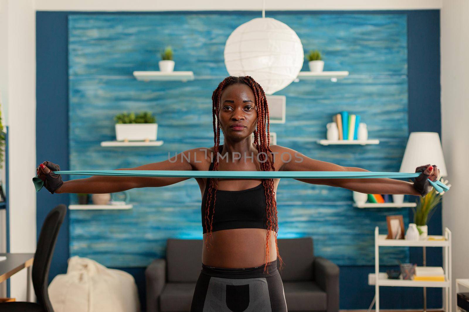 Black strong fit woman standing in living room working out shoulders using resistance band dressed in sportive sportwear. Active african exercising with rubber band.