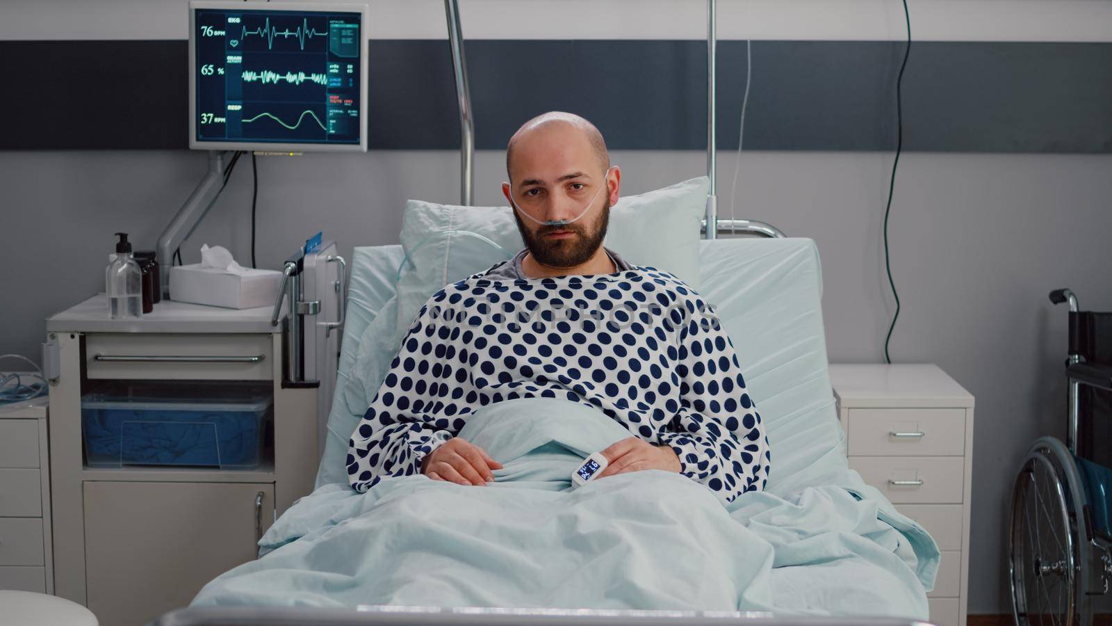 Potrait of sick man resting in bed waiting for respiratory treatment recovering after medical surgery in hospital ward. Hospitalized patient looking into camera wearing nasal oxygen tube