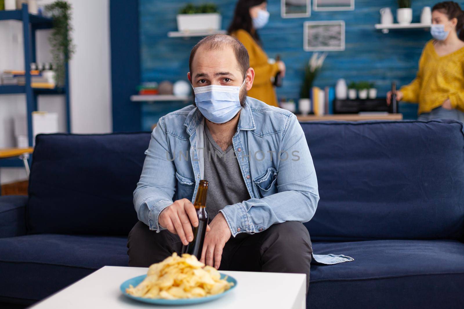 Man holding beer bottle relaxing on couch spending time with friends keeping social distancing with face mask in living room looking pensive at camera preventing spread of coronavirus.