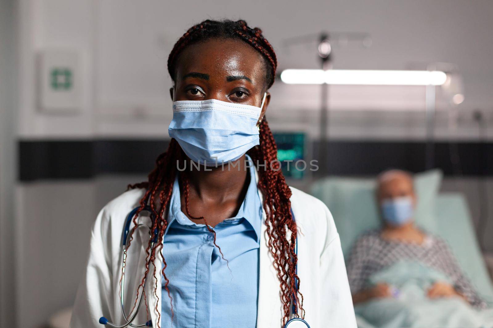 African american doctor health care specialist wearing chirurgial mask in hospital room with sick patient btreathing with help from oxygen tube and iv drip attached.