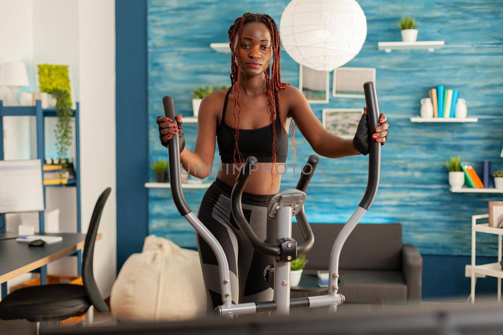 Black person training in home living room using eliptical machine for cross cardio training, watching online exercising. Sporty strong active woman with healthy lifestyle.