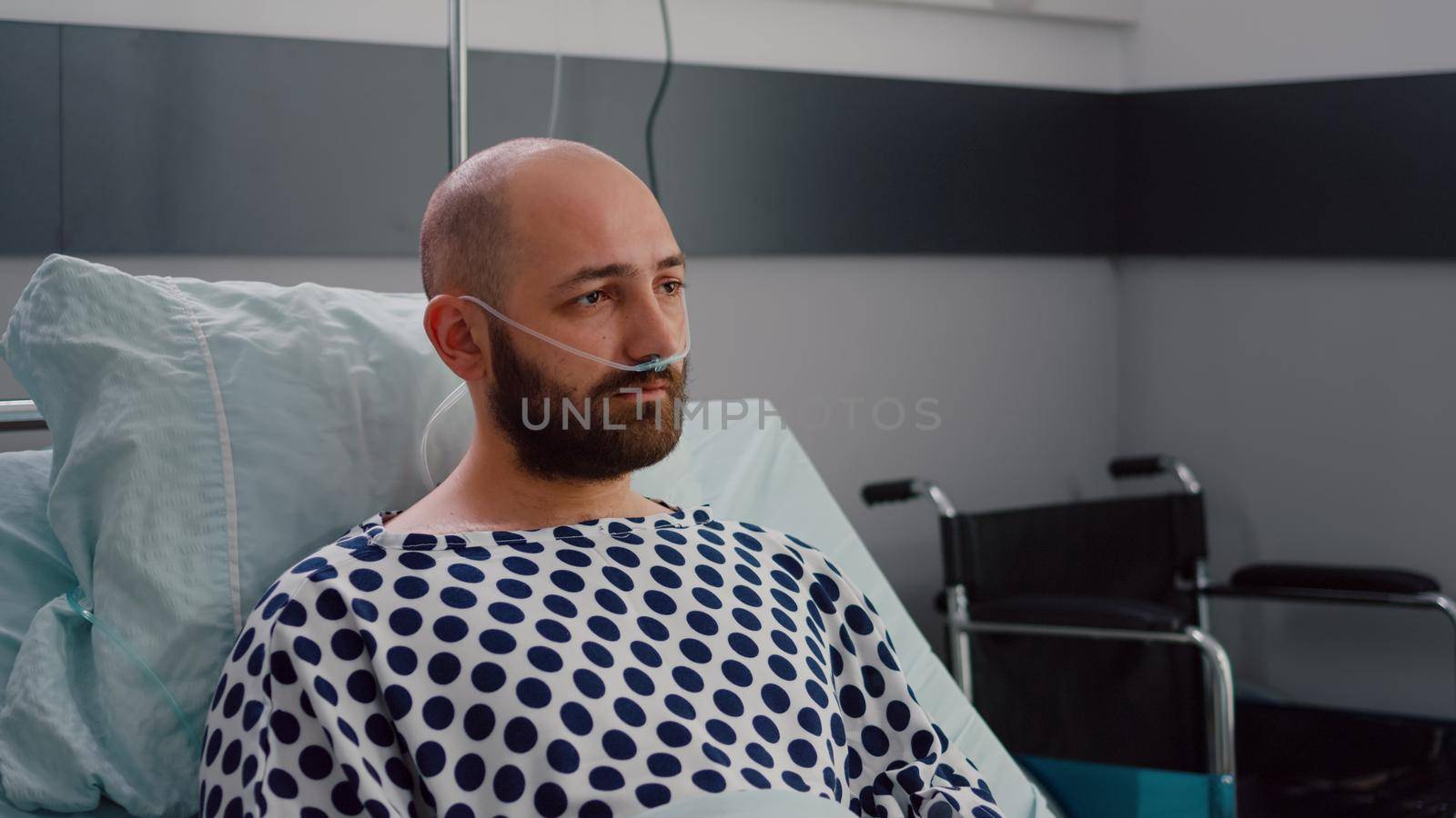 Sad sick man with nasal oxygen tube resting in bad looking into camera during respiratory recovering in hospital ward. Depressed patient waiting for illness breathing treatment