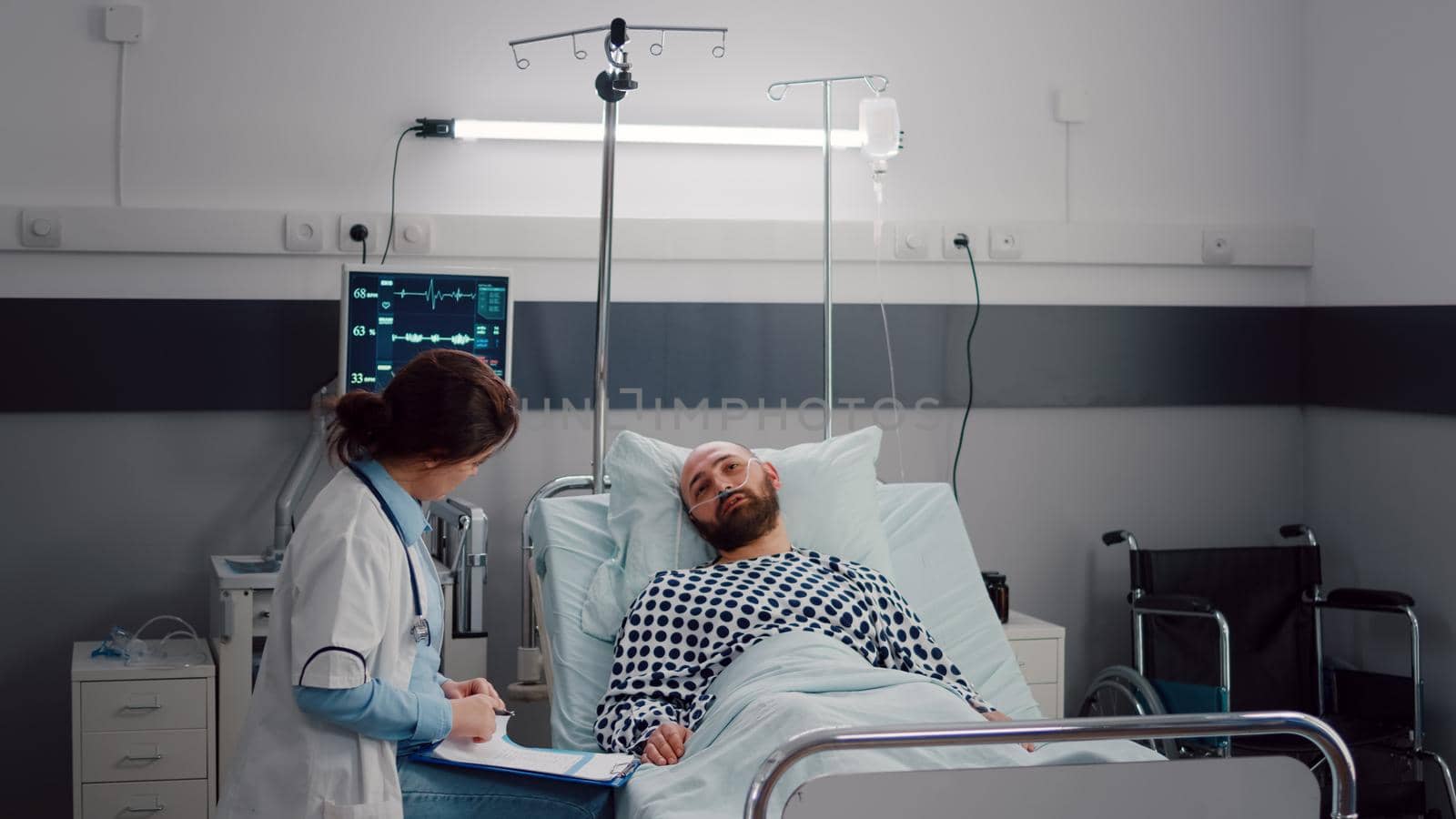 Practitioner doctor monitoring sick man writing disease treatment on clipboard checking sickness symptom. Medical nurse arrange patient bed in hospital ward during recovery appointment