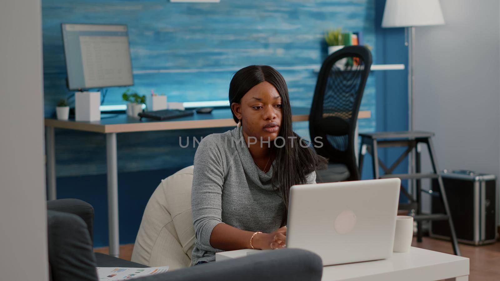 Blogger student with dark skin working from home browsing communication information while drinking coffee sitting on bean bag in living room. African woman working from home at media webinar