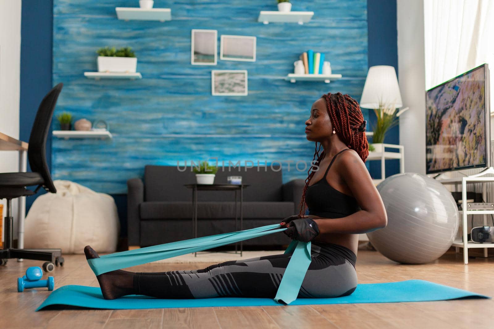 Fit strong afro woman training home living room using resistance band, sitting on fitness mat pulling for back muscles. Training using arm rowing movement holding rubber band.