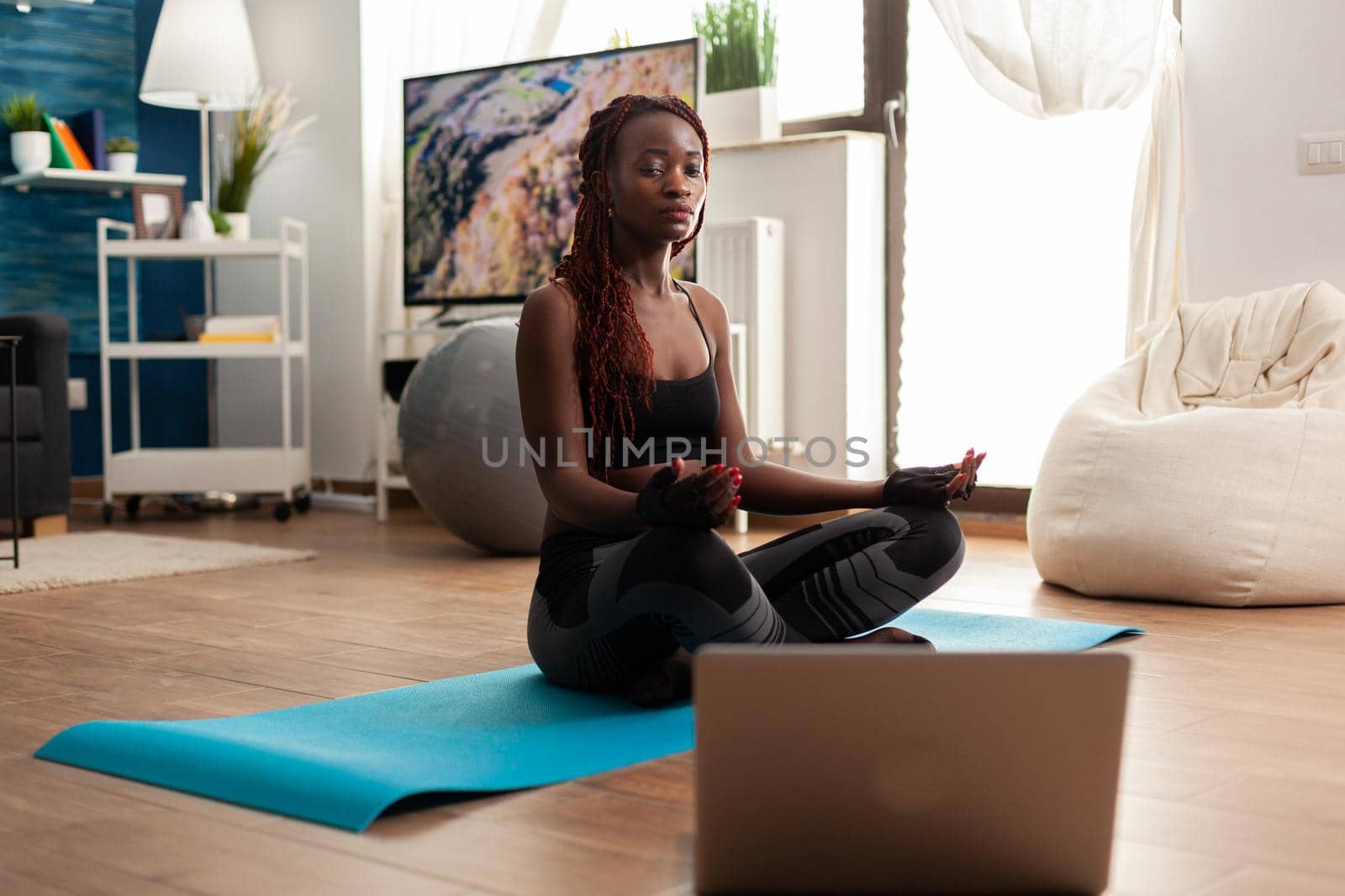 Black woman sitting on yoga mat practicing calm harmony mediting zen for healthy lifestyle, relaxing in lotus pose. Listening instructor instruction during online training.