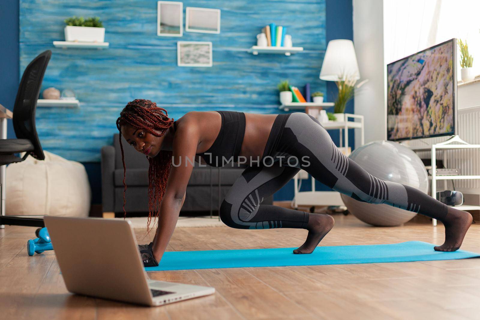 Black fit athletic woman training for muscle strenght doing mountain climbers position on yoga mat dressed in sportwear leggings, in home living room following online instructions.