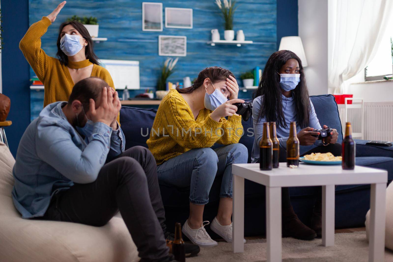 Sad multi ethnic group of friends after lose at gaming competiton with face covers with mask during covid outbreak keepin social distancing in home living room. Enjoying beer and chips. Conceptual image.