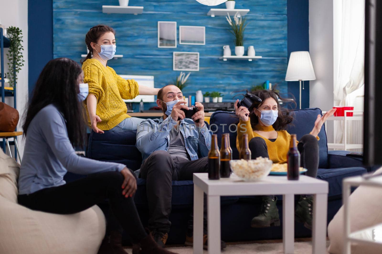 Excited group of multi ethnic friends scraming celebrating victory during gaming competition wearing face mask keeping social distancing to prevent coronavirus spread in time of global pandemic.