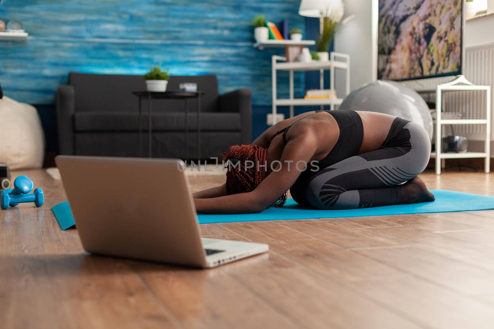 Black woman sitting on yoga mate doing stretching relaxing body after watching online training looking at laptop in home living room, on the floor for health wellbeing.