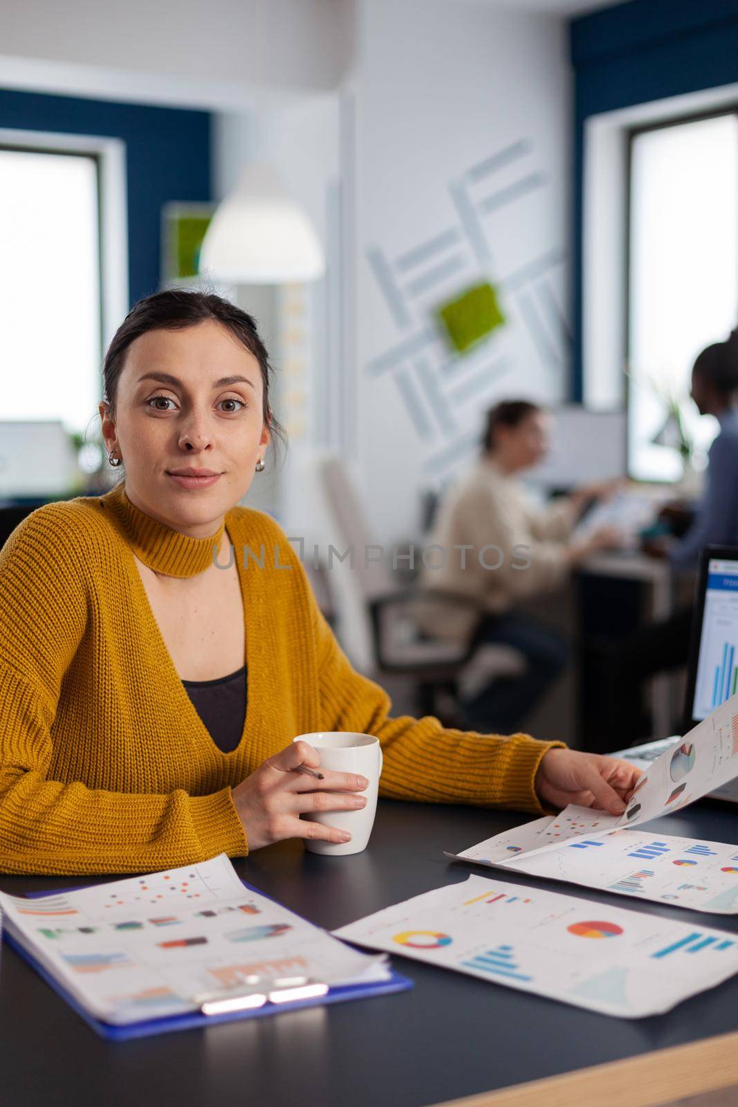 Businesswoman working with company charts looking at camera holding cup of coffee. Executive entrepreneur, manager leader sitting working on projects with diverse colleagues.