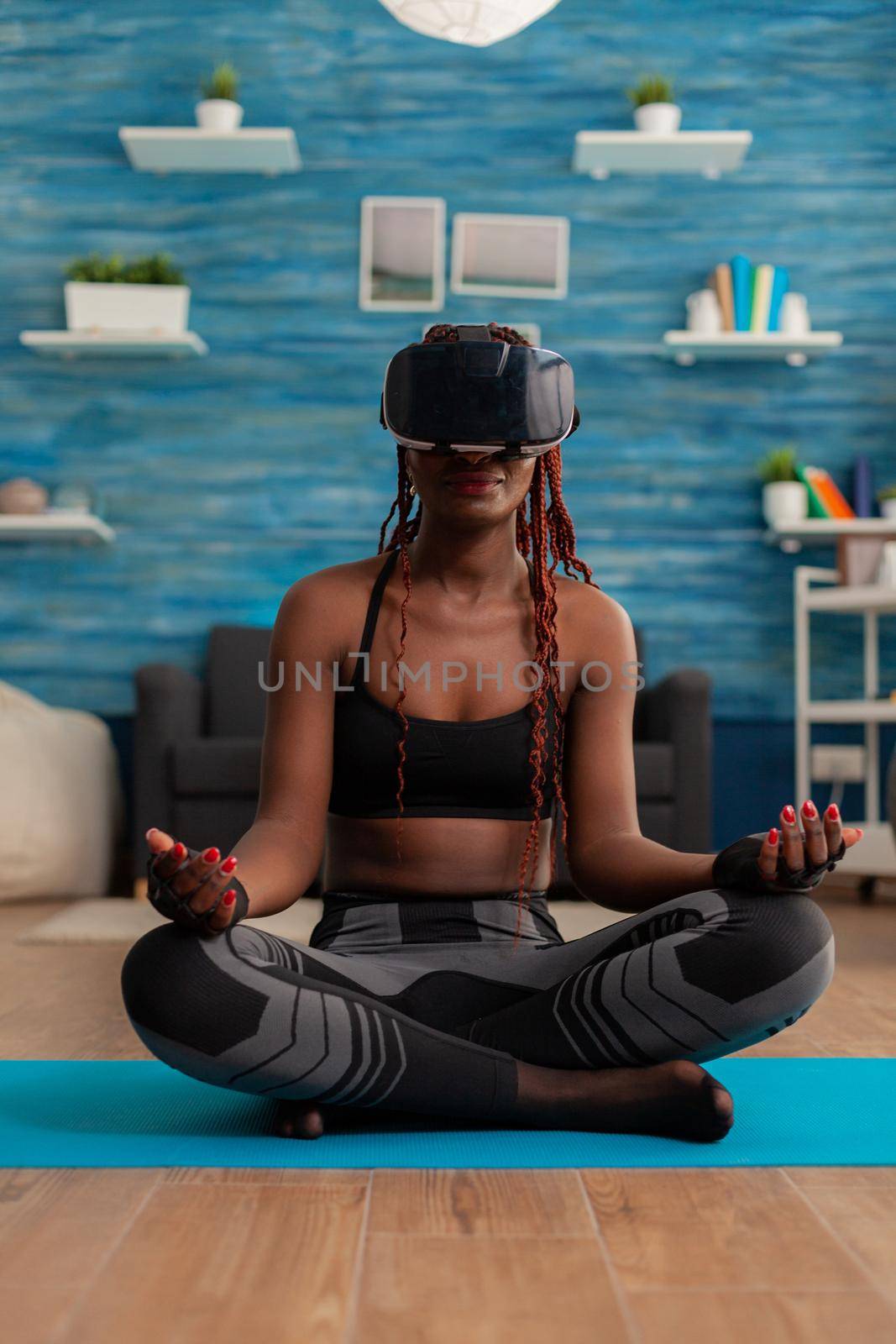 Calm tranquil black woman wearing vritual reality goggles sitting on yoga mat, spending time on spirituality healthy lifestyle. Practicing meditating in lotus pose in home living room.