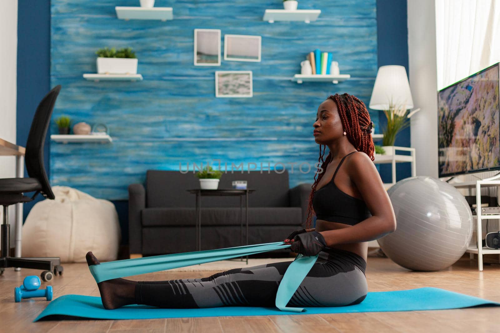 Black woman doing pilates workout using elastic strap sitting on yoga mat, pulling training arms and shoulders in home living room. Athletic fit exercising body using resistance band.