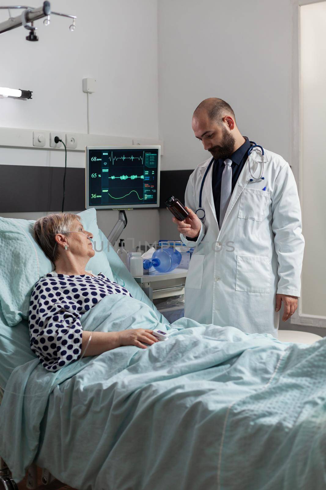 Senior woman patient breathing with help from oxygen tube laying in hospital bed, listening doctor explaying diagnosis treatmant holding pills bottle for recovery.