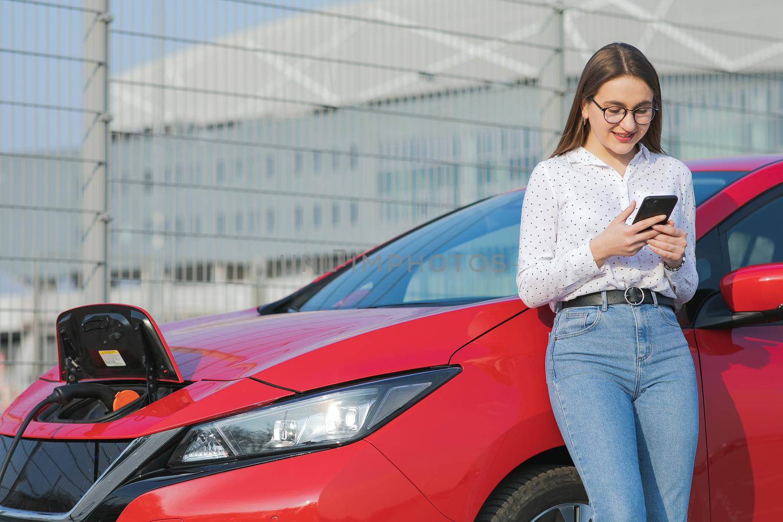 Woman stands with phone near her red electric car and waits when vehicle will charged