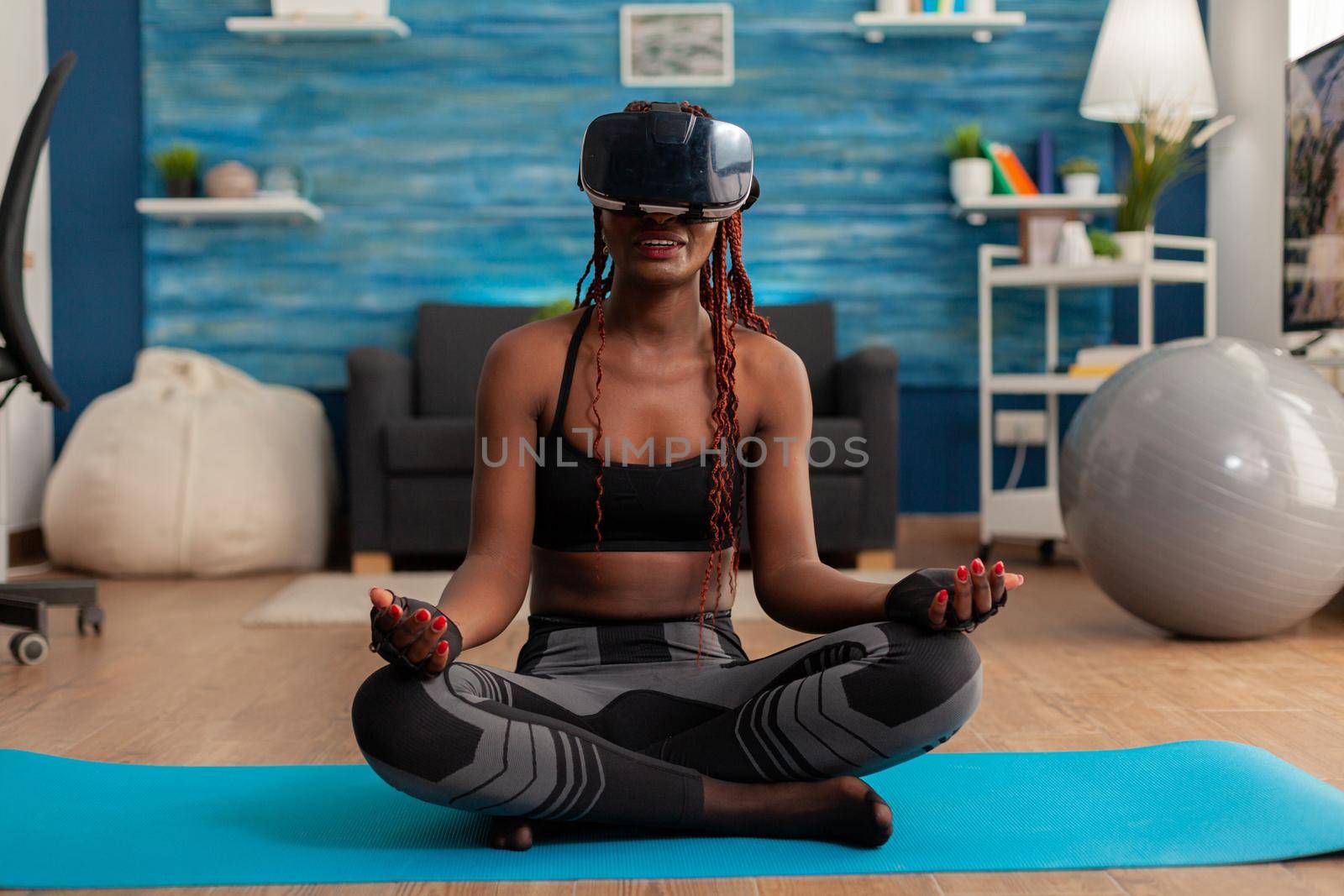 Black woman experiencing virtual reality training body and mind meditating in lotus pose sitting on yoga mat in home living room for calm, healthy harmony lifestyle dressed in sportwear.