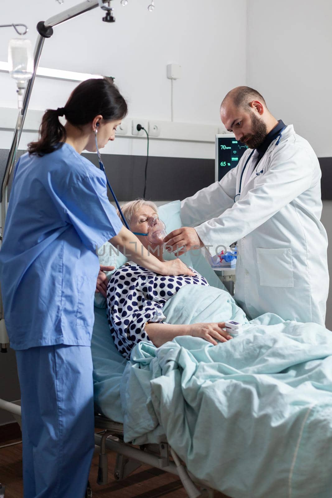 Doctor holding oxygen mask for senior woman patient helping her breath, in hospital room with respiratory failure. Nurse using stethoscope listening sick elderly heart in clinic. Iv drip bag attached.