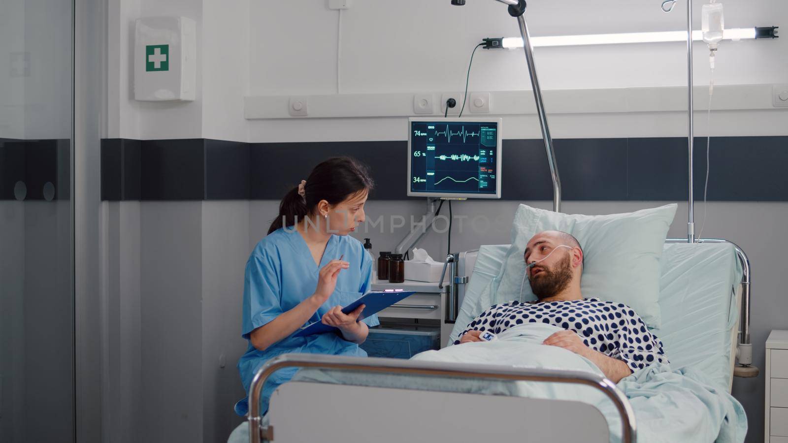 Medical nurse discussing illness treatment with hospitalized sick man resting in bed during rehabilitation therapy in hospital ward. Patient with nasal oxygen tube explaining disease symptom