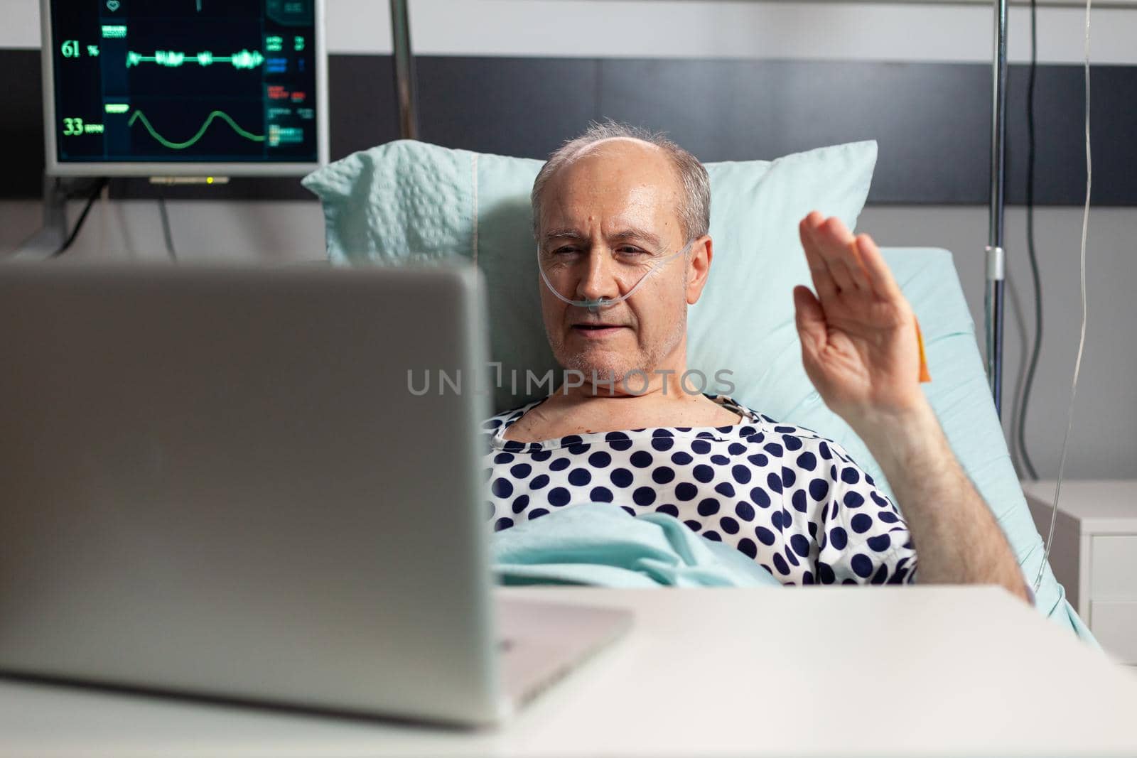 Cheerful sick senior man waving at camera during video conference using laptop laying in bed, breathing through oxygen tube. Man recovering after surgery and treatment.