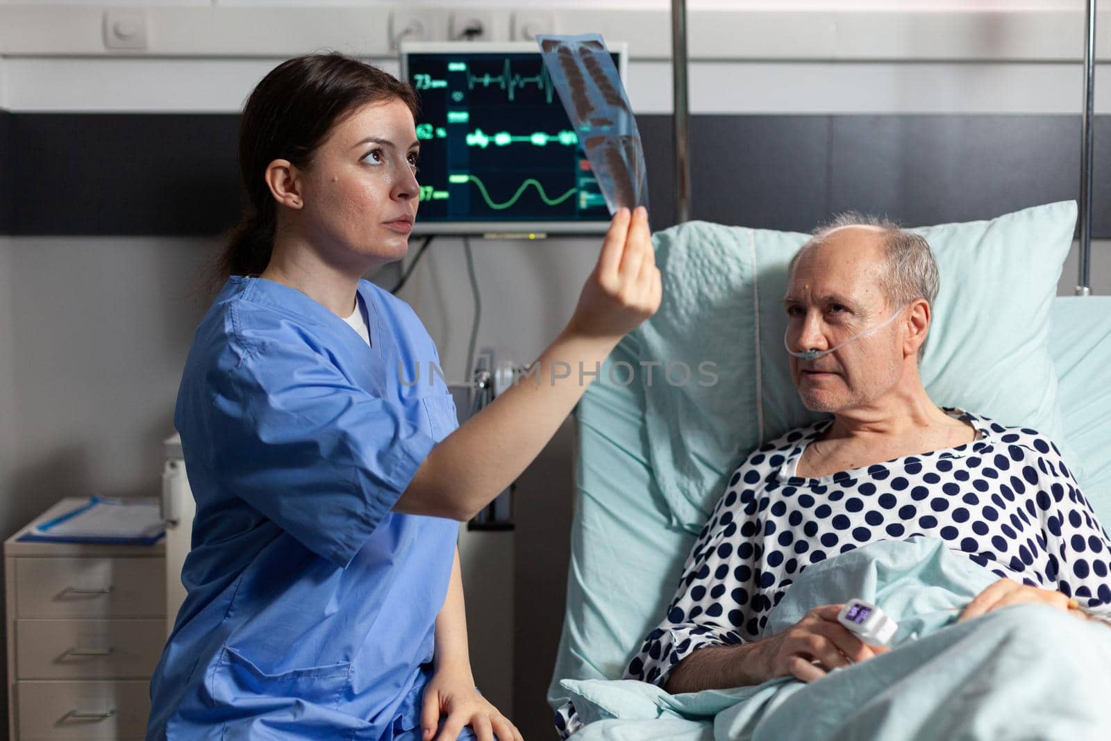 Medical nurse analyzing senior patient chest x-ray in hospital room, discussing explaining diagnosis. Old man breathing through respiratory tube laying in bed listening health care specialist.