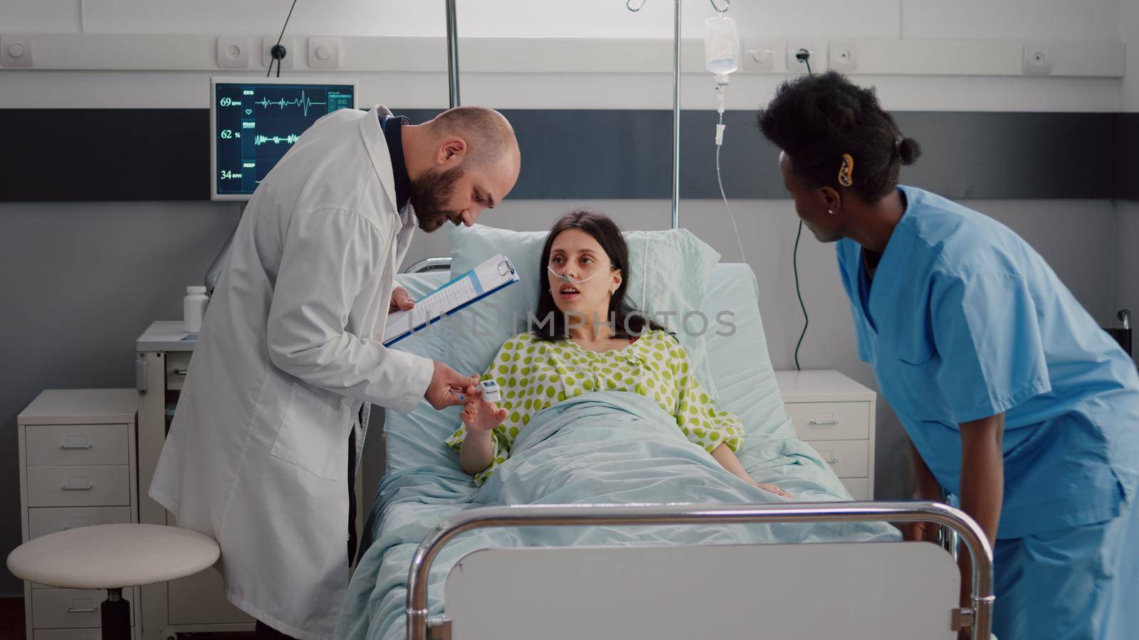 Specialist doctor monitoring sick woman examining pulse oximeter writing disease treatment on clipboard. Patient resting in bed while black nurse make bed comfortable in hospital ward