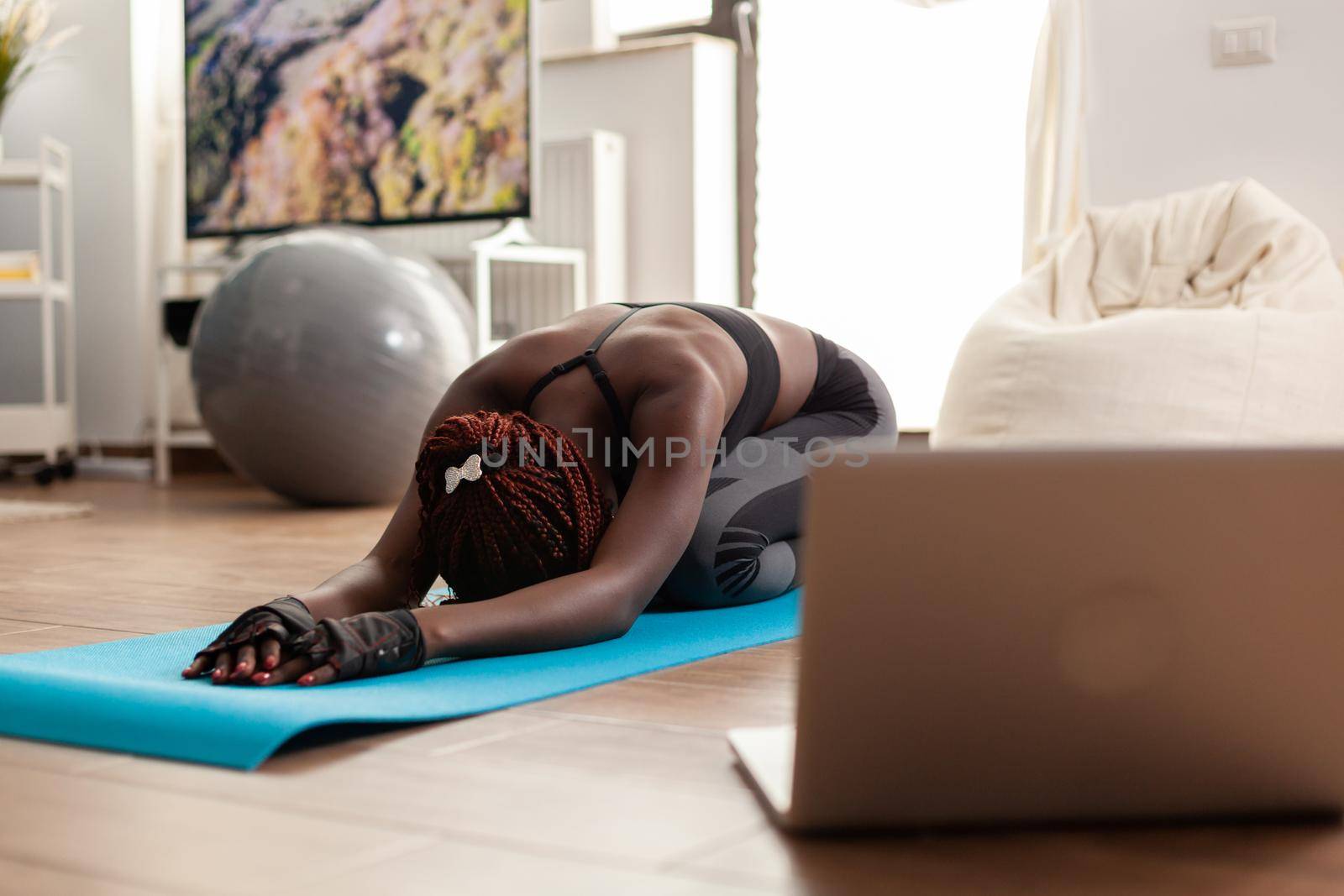 Black woman exercising and stretching her body doing flexibility exercises sitting on yoga mat in home living room wearing leggings during online tutorial. Workout and healthy lifestyle.