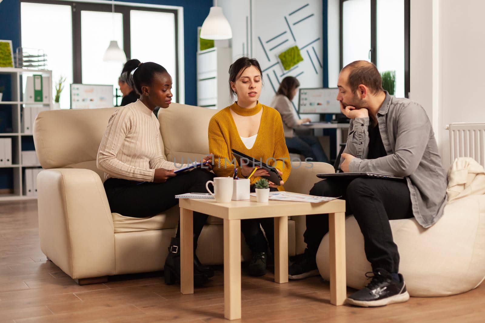 Concentrated multiethnic businesspeople reading annual financial report from tablet sitting on couch in modern startup business office. Diverse team of multiethnic businesspeople working in company