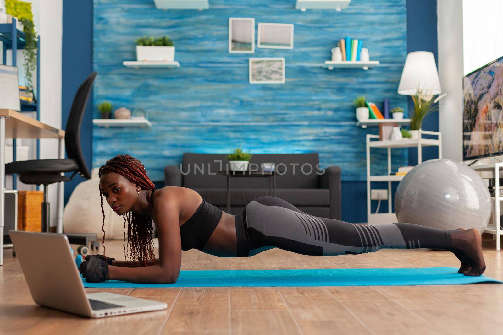 Athletic fit black woman in leggings sitting on yoga mat doing plank pose, exercising core abdominal strenght for muscle balance watching online training in home living room.