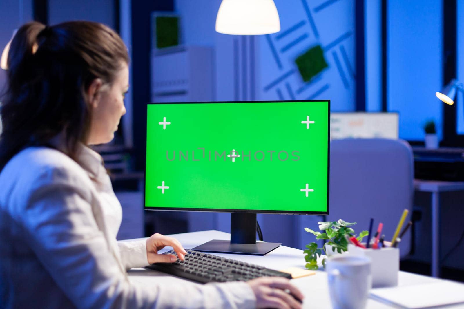 Entrepreneur working in front of green screen display sitting at desk during videocall meeting in business office. Businesswoman watching desktop monitor with green mockup, chroma key, working overtime.