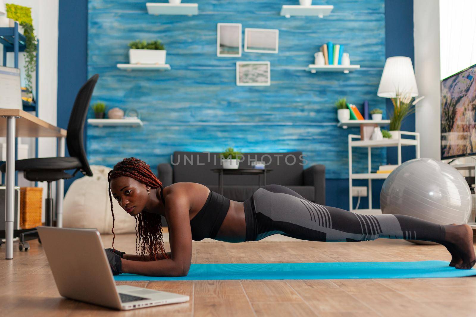 Athletic fit black woman in leggings sitting on yoga mat doing plank pose, exercising core abdominal strenght for muscle balance watching online training in home living room.