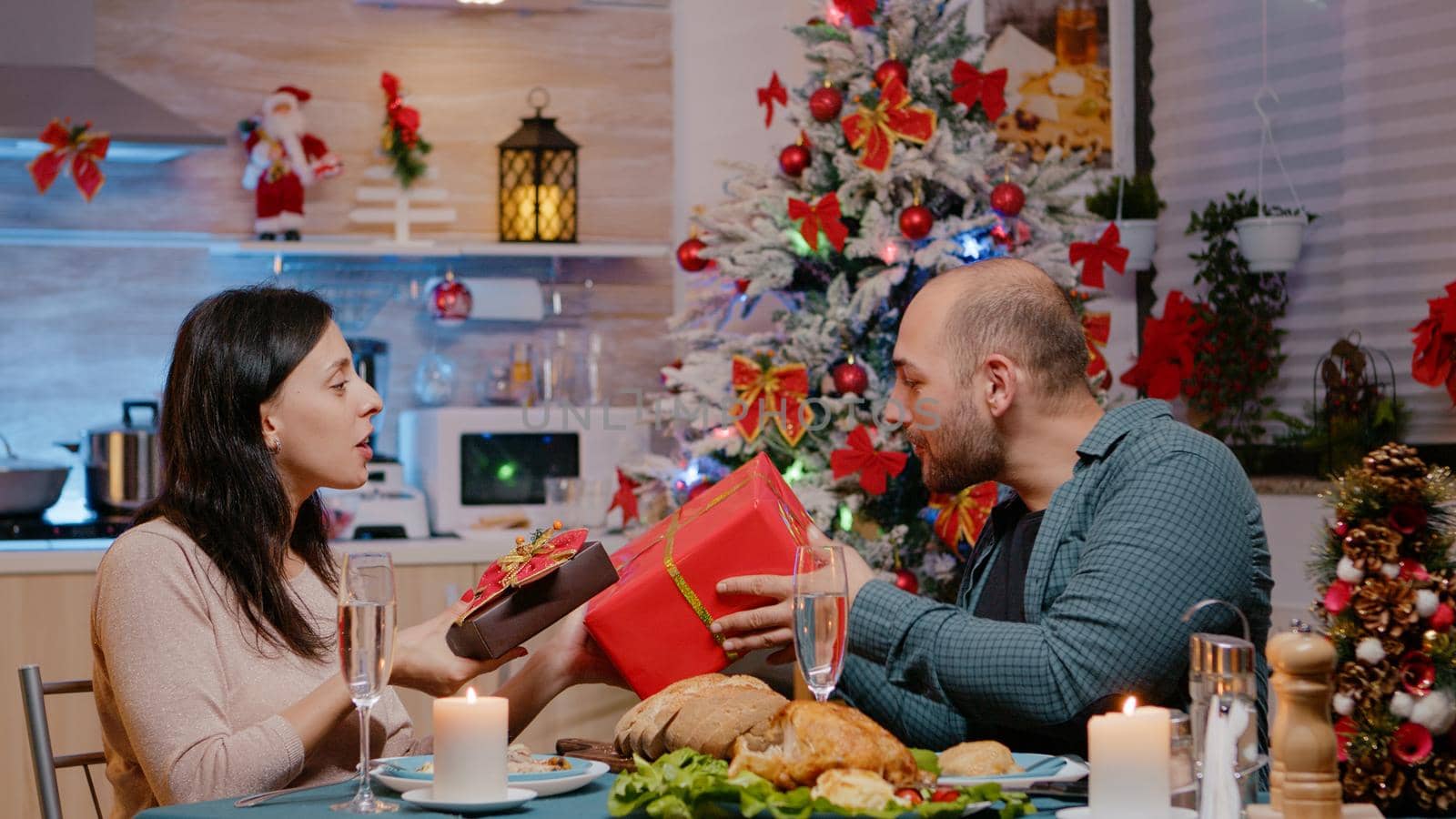 Festive couple giving and receiving gifts at christmas dinner at celebration. Man and woman celebrating traditional holiday, having presents and seasonal meal with glasses of champagne.