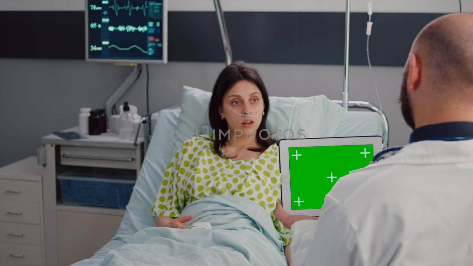 Specialist man in white coat talking with sick woman about illness recovery treatment. Practitioner holding mock up green screen chroma key tablet computer with isolated display in hospital ward