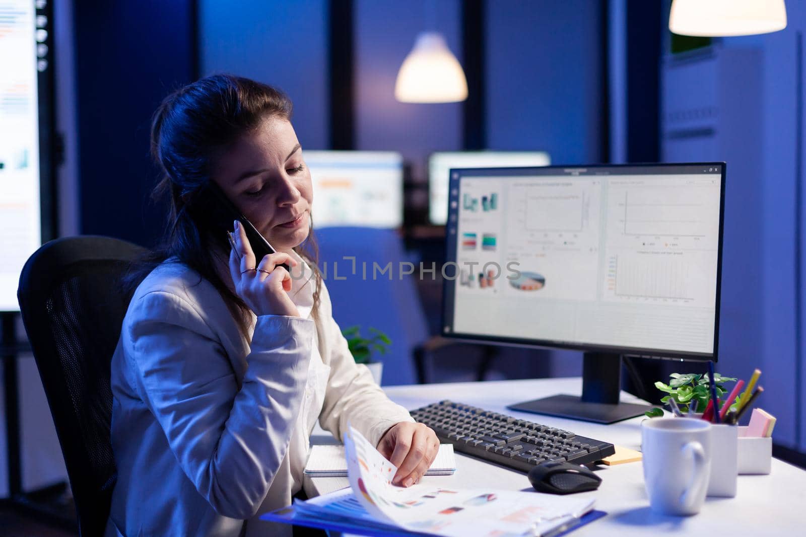 Executive entrepreneur talking at smartphone with employee creates new marketing concept in business office. Busy manager using modern technology network sitting at desk late at night